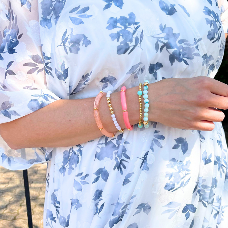 Model wearing 5-peice beach inspired stack. Designed with blue opal gemstone beads with gold filled round beads. Two acrylic 8mm skinny tube bangles with gold plated flats. Acrylic tubes are peach marble and watermelon. The other two bracelets in this set are dainty and are adorned with gold filled round beads. 6mm blue acrylic beads and 5mm white acrylic beads accent the gold bracelets.