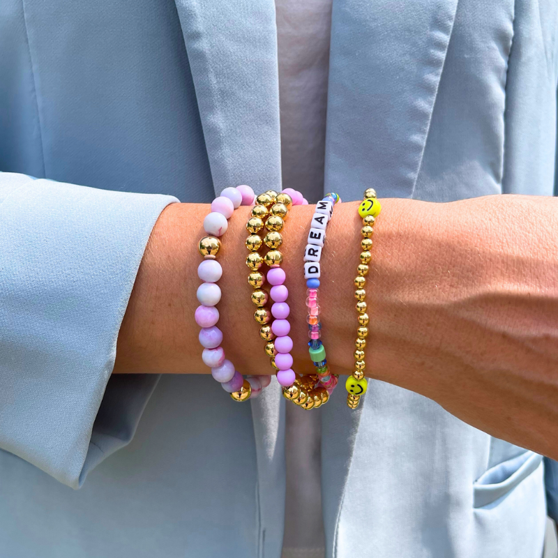 Model wearing 5 bracelets from the Turks and Caicos stack. Purple opal gemstones adorn one bracelet. 2 of the bracelets are designed with 4mm gold filled round beads and one has yellow smiley face beads. Another bracelet has light purple acrylic beads with gold filled round beads. Add a personal touch with a dainty rainbow beaded word bracelet with 4mm white cubed letter beads.