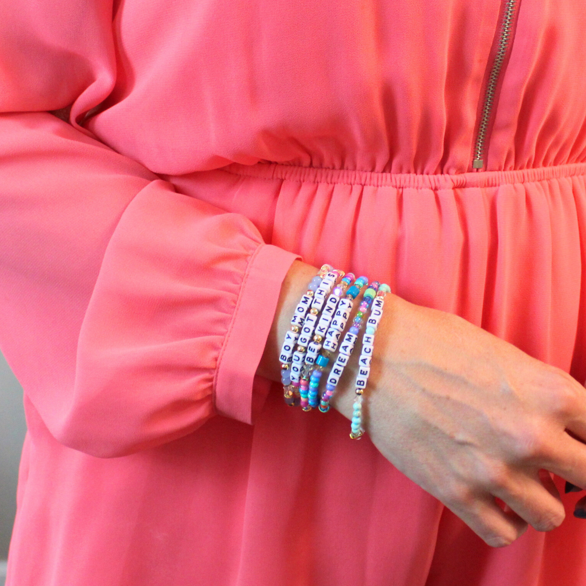 Model wearing stacks of different letter bead bracelet designs. Each bracelet is designed with 5mm white cubed letter beads with black letters. Bracelets are full of bold colors and can be personalized.