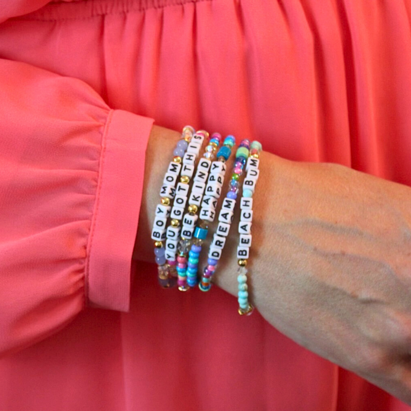 Model wearing stacks of different letter bead bracelet designs. Each bracelet is designed with 5mm white cubed letter beads with black letters. Bracelets are full of bold colors and can be personalized.