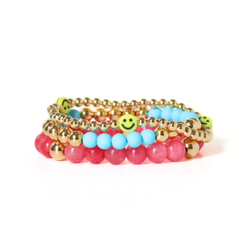Picture shows a 3-piece bracelet stack flat. This bracelet set has a dainty 18k gold filled beaded bracelet with 3 bright yellow smiley face bracelet. Also designed with blue acrylic beads with 18k gold filled beads. The third bracelet is a pink gemstone beaded bracelet with 3 gold filled round beads.