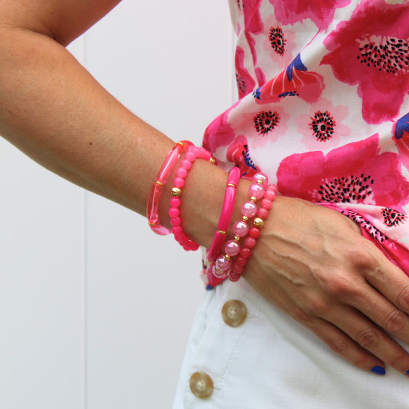 Model wearing the Tahiti bracelet stack. This 5-piece bracelet stack has 5 pink acrylic bracelets. The acrylic bracelets are different shades of pink and textures. 2 bracelets are pink acrylic tubed beads with gold flat beads. 2 bracelets are acrylic round beads with gold fille round beads. The last bracelet has chunky pink iridescent wound beads.