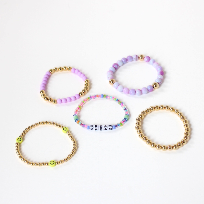 5 bracelets from the Turks and Caicos stack laying flat. Purple opal gemstones adorn one bracelet. 2 of the bracelets are designed with 4mm gold filled round beads and one has yellow smiley face beads. Another bracelet has light purple acrylic beads with gold filled round beads. Add a personal touch with a dainty rainbow beaded word bracelet with 4mm white cubed letter beads.
