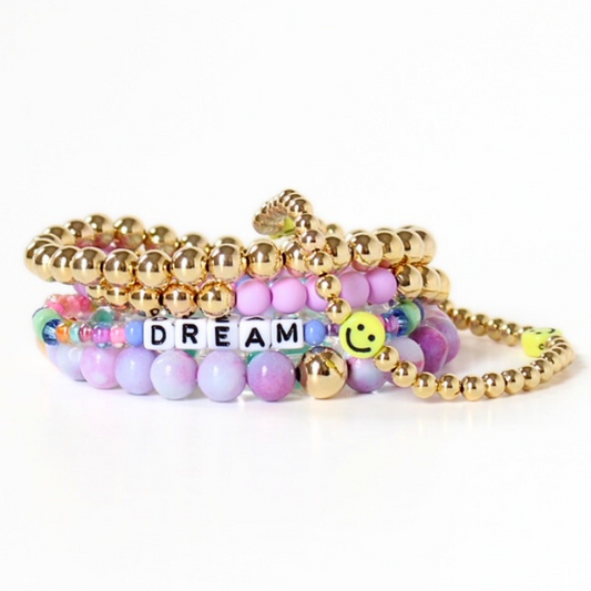 Stack of 5 stretch bracelets designed with purple acrylic beads and purple opal gemstones. This bracelets is adorned with 18k gold filled round beads. Adding a playful look a letter bead bracelet and a smiley face beaded bracelet add a personal touch.