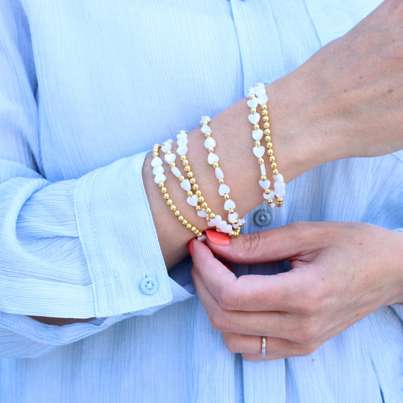 Model wearing stack of two different styles of 14k gold filled beaded stretch bracelet with mother of pearl heart charms. Design one has alternating 4mm gold beads and mother of pearl heart charms. The second bracelet has three mother of pearl heart charms followed by gold beads.